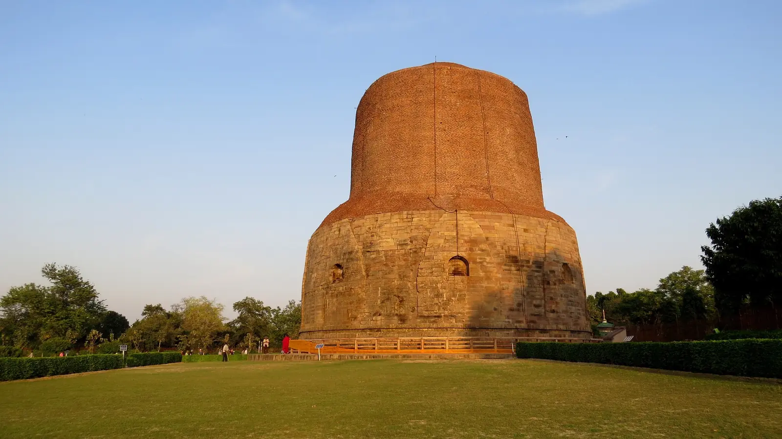 The massive cylindrical brick and stone Dhamek Stupa at Sarnath in warm evening light, with lawns and trees around it