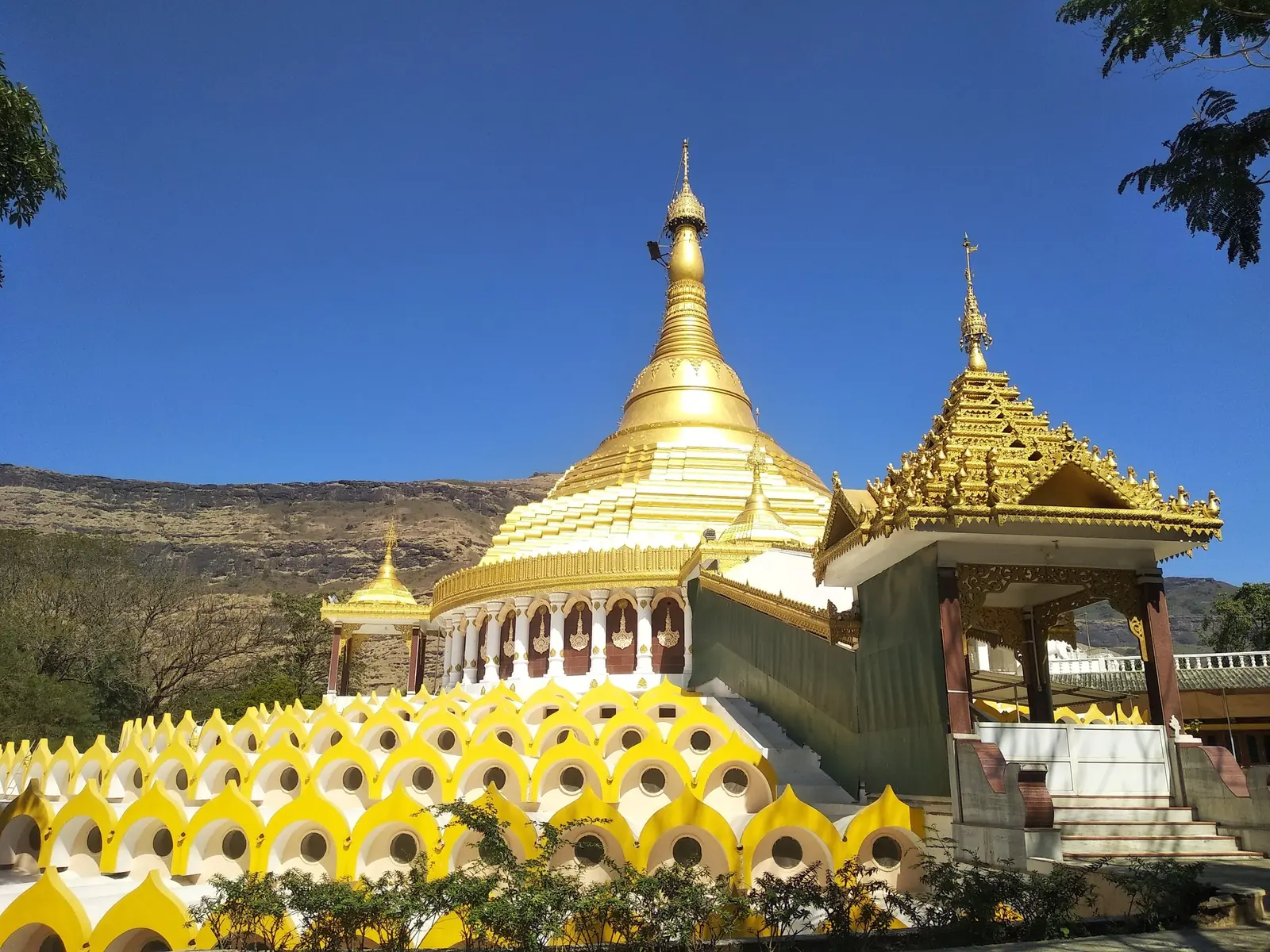 The golden pagoda at Dhamma Giri, Igatpuri, behind a scalloped yellow-and-white ornamental wall, with a gilded Burmese-style pavilion and dry hills beyond