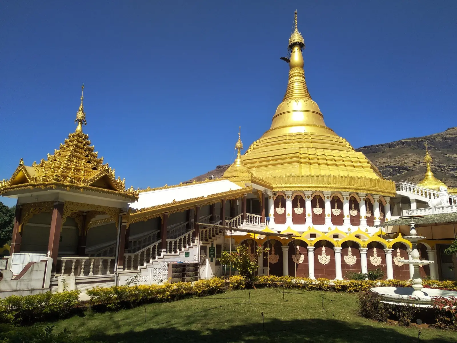 The Dhamma Giri pagoda with its arcaded base, a gilded Burmese-style pavilion and a lawn with a fountain, under a deep blue sky