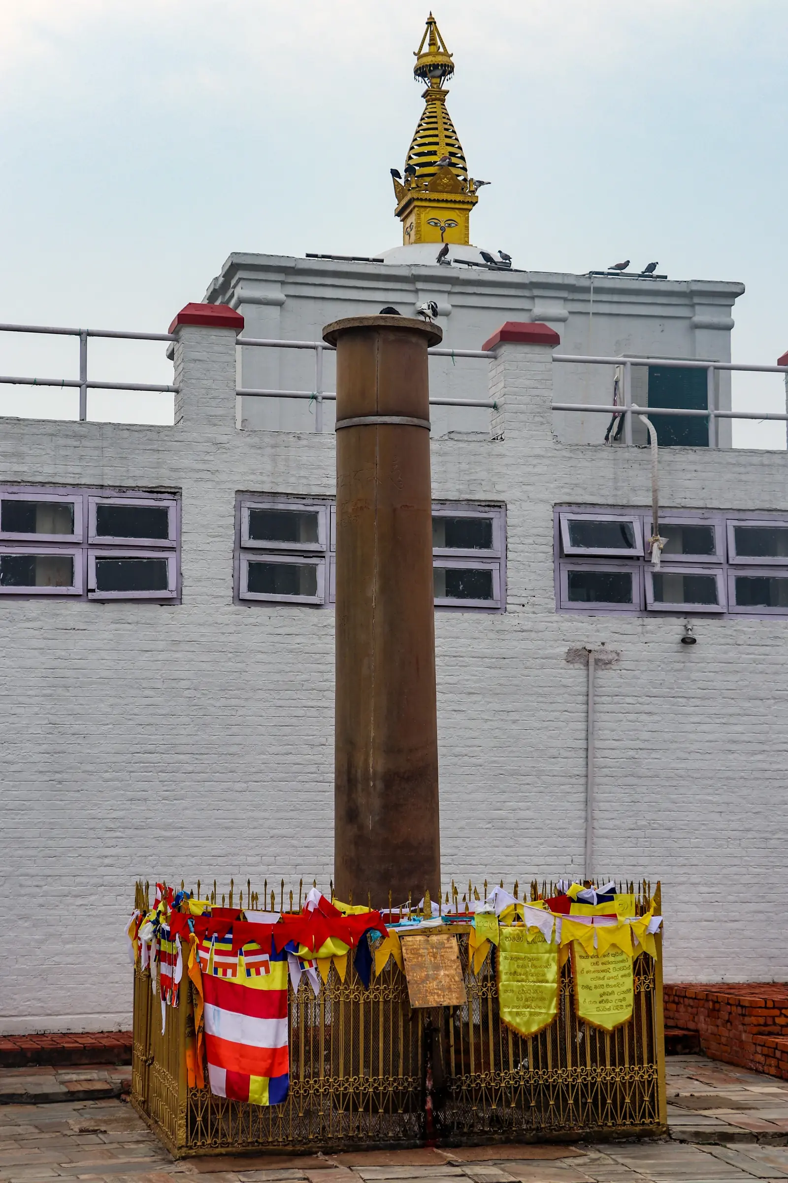 Close view of the Ashoka pillar at Lumbini, its railing hung with prayer scarves, with the Maya Devi Temple behind