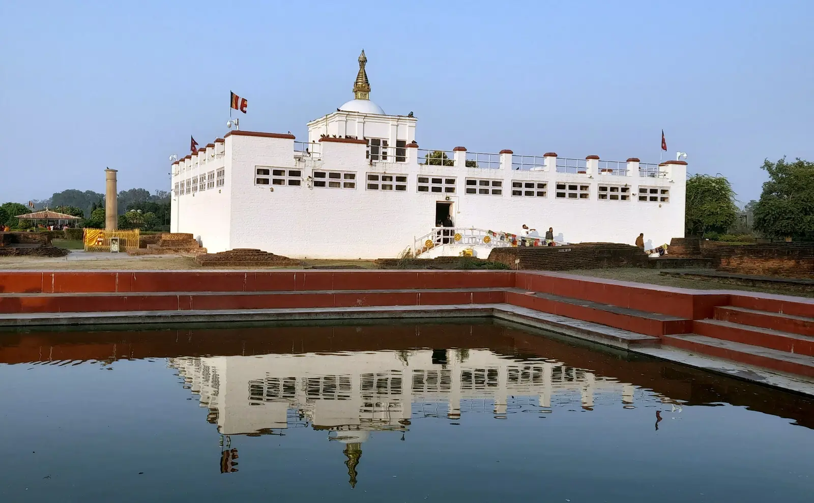The white Maya Devi Temple at Lumbini reflected in the still water of the brick-lined sacred pond, with the Ashoka pillar at left