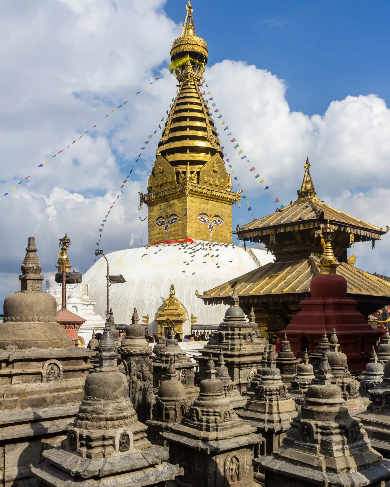 The gilded spire of Swayambhunath Stupa with the painted Buddha eyes, above clusters of small stone votive stupas under white clouds