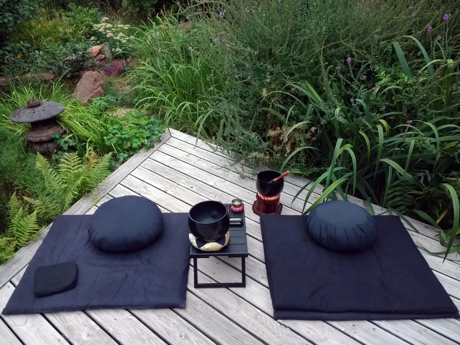 Two round dark-blue meditation cushions on square mats laid out on a wooden deck beside a garden, with a small table holding a bowl-bell