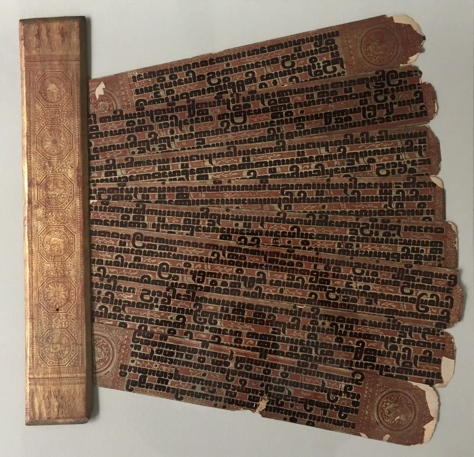 Leaves of a 19th-century Burmese Pali manuscript, lacquered and gilded palm leaves written in bold black square script, beside its gilded wooden cover