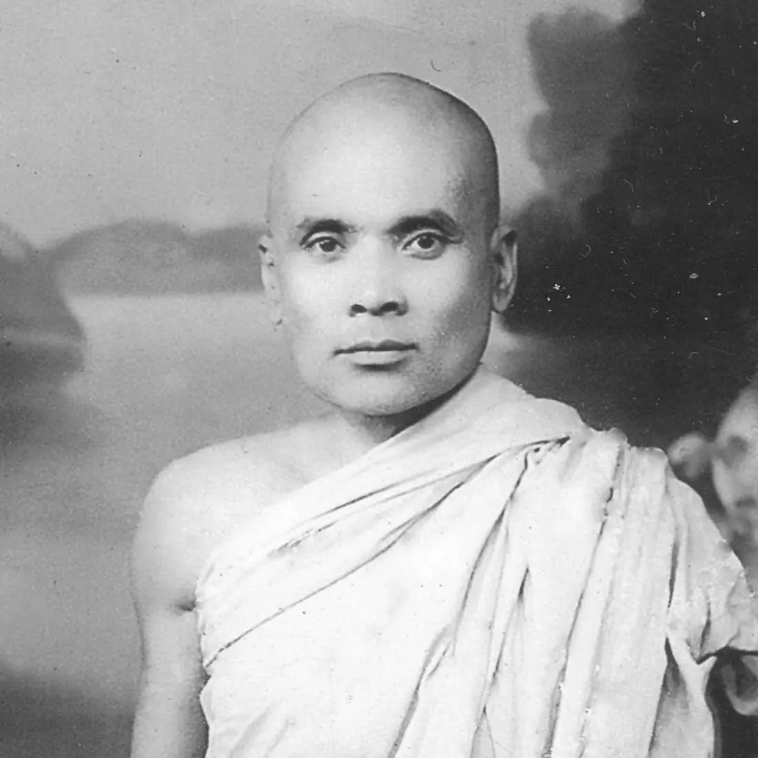 Black-and-white studio portrait of Bhikkhu Dhammalok, a shaven-headed monk in a pale robe worn over one shoulder, in front of a painted landscape backdrop