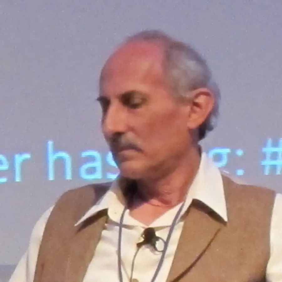 Jack Kornfield, grey-haired with a moustache, in a white shirt and tan waistcoat, looking down while seated on a conference stage