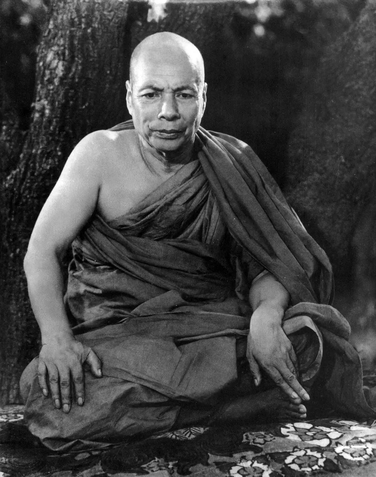 Black-and-white portrait of Mogok Sayadaw seated cross-legged on a patterned mat in front of a tree, one shoulder bare