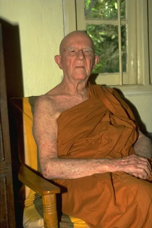 Nyanaponika Thera, an elderly monk in orange robes, seated in a wooden armchair beside a window