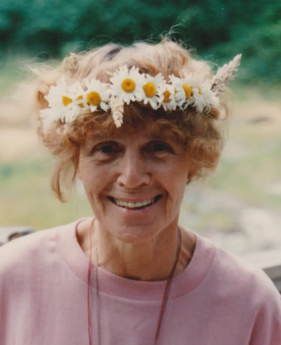 Ruth Denison smiling outdoors, wearing a crown of daisies and a pink top