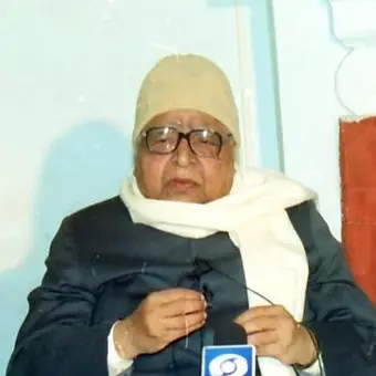 S. N. Goenka seated indoors, wearing glasses, a cream knitted cap, a white scarf and a dark coat, with a news microphone in front of him