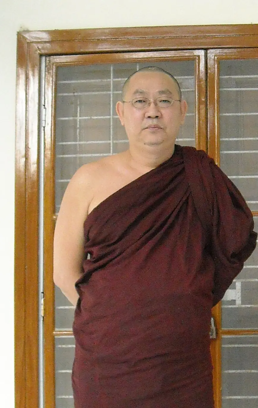 Sayadaw U Tejaniya, a Burmese monk in glasses and maroon robes, standing with his hands behind his back in front of a wooden door